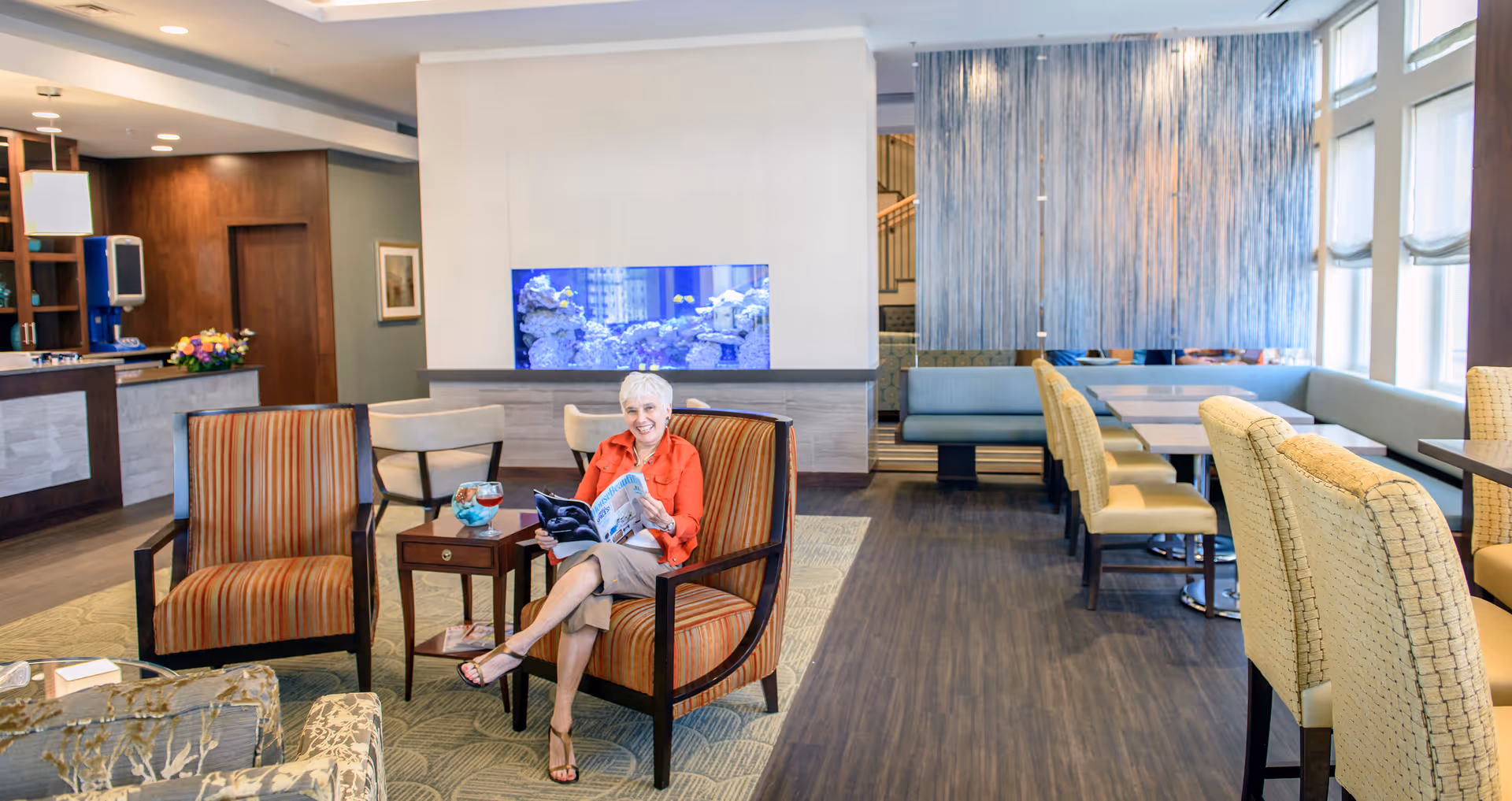 Bright senior living common area with lounge chairs, dining tables, an aquarium, and a person reading a magazine.