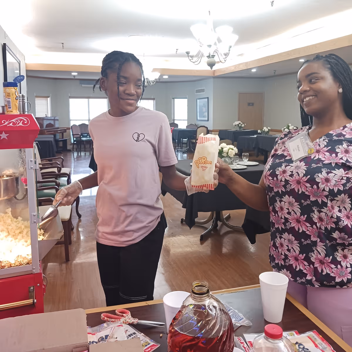 Two women smiling and interacting near a popcorn machine in a dining area. One woman is scooping popcorn while the other is holding a popcorn bag. The room has tables with black tablecloths and floral centerpieces.