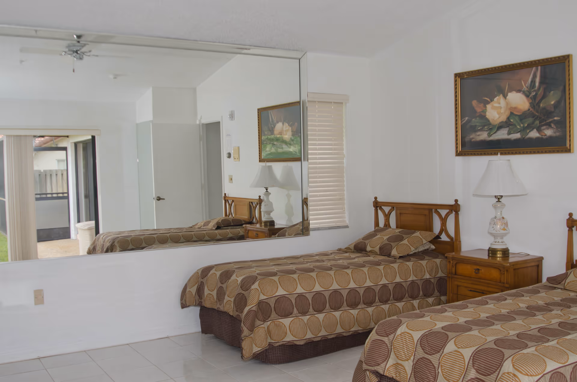 A bedroom with two single beds covered in brown and beige patterned bedspreads. Between the beds is a wooden nightstand with a decorative lamp. A large mirror covers most of one wall, reflecting the beds and part of the room. A framed floral painting hangs above the nightstand, and a window with closed blinds is visible on the adjacent wall. The floor is tiled in white.
