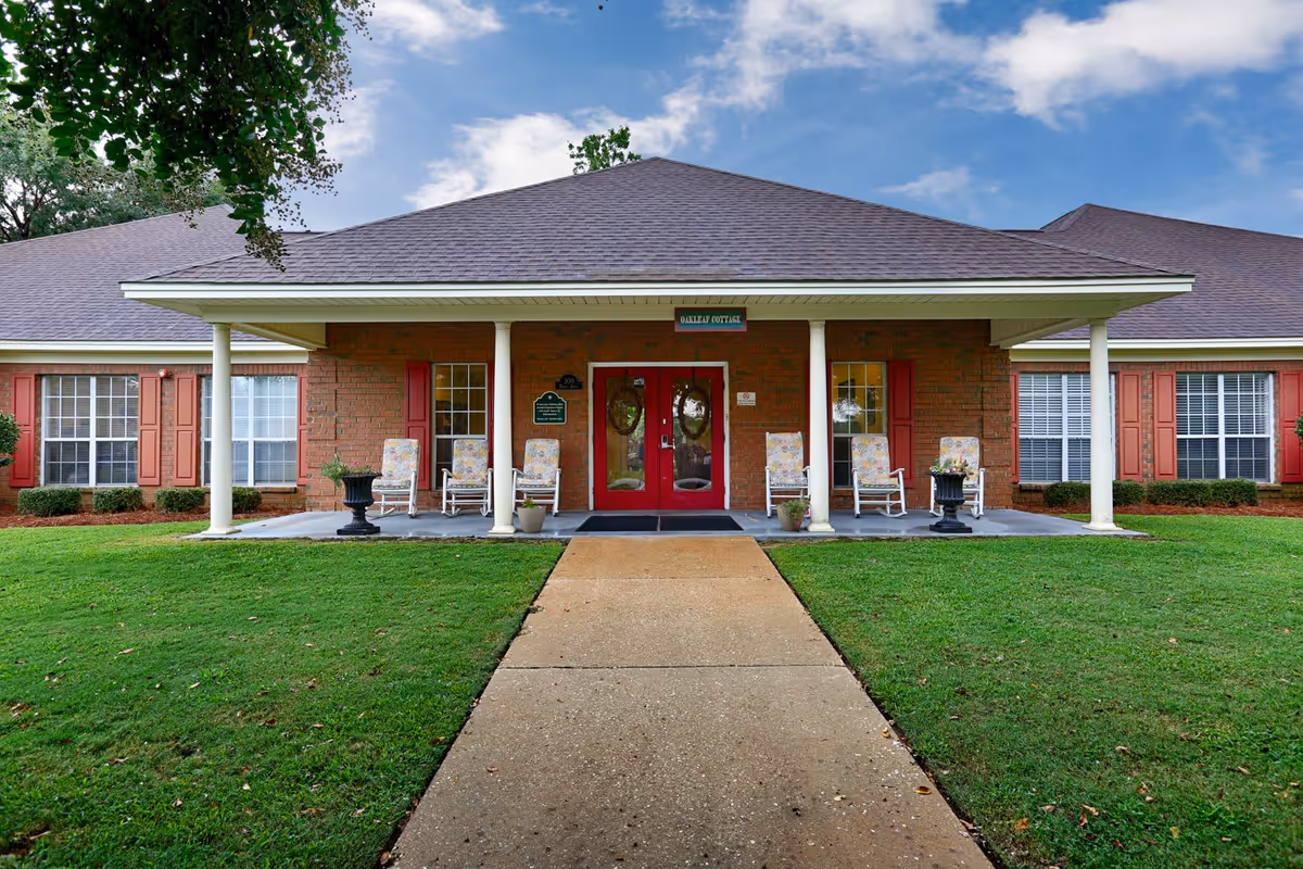 Front exterior view of a single-story brick building with a covered porch supported by white columns. The porch has several cushioned rocking chairs and potted plants. The building has red-framed double glass doors and multiple windows with red shutters. A concrete pathway leads up to the entrance, and the surrounding area is a well-maintained green lawn with some trees and shrubs.