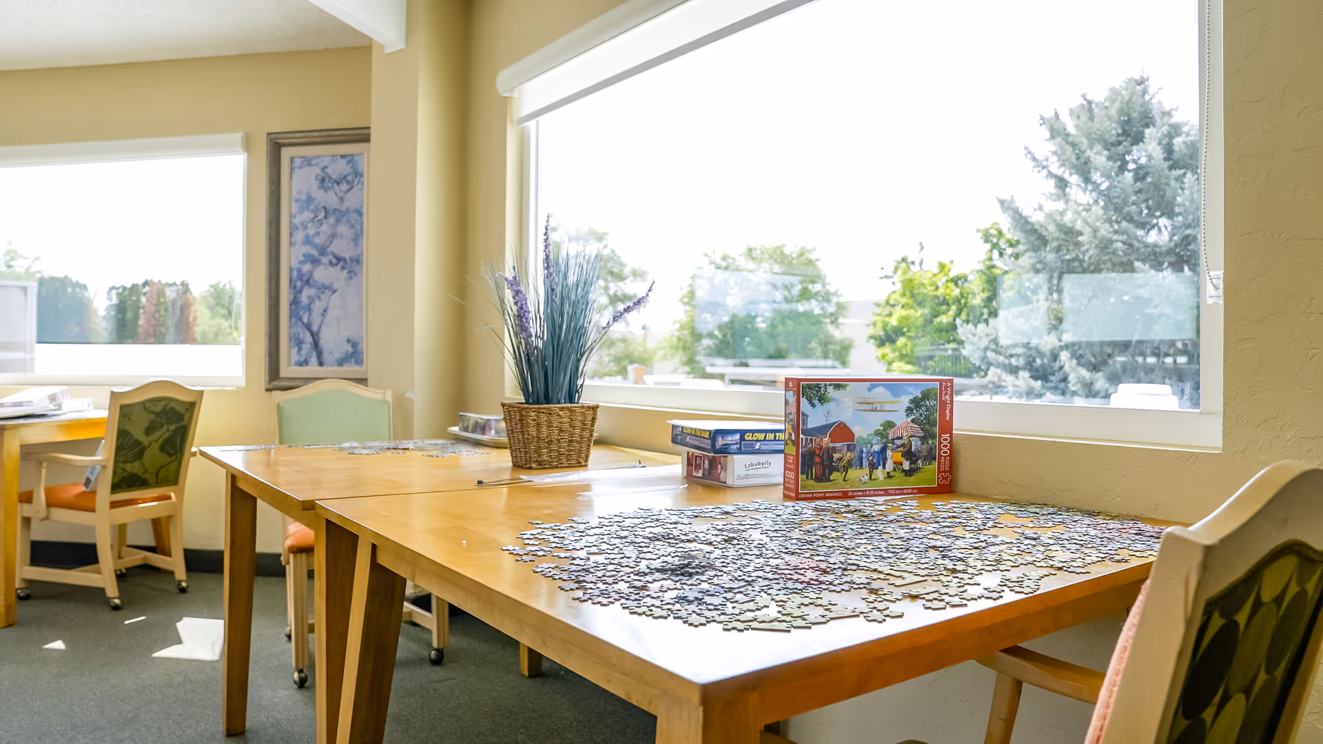A bright room with large windows showing greenery outside. Inside, there are wooden tables with chairs around them. On one table, there is a partially completed jigsaw puzzle with puzzle boxes stacked nearby and a basket with decorative plants.