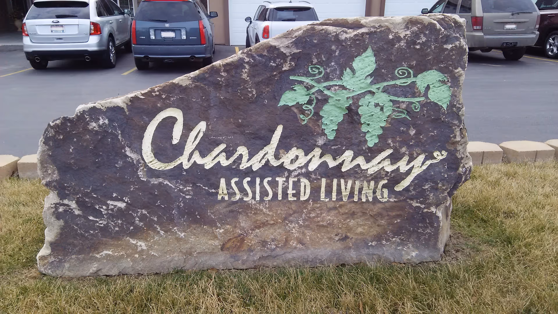 A large decorative rock with the words 'Chardonnay Assisted Living' painted on it, along with a green grapevine design. The rock is situated on grass in front of a parking lot with several parked cars.