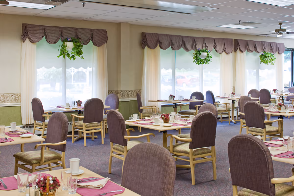 A bright dining room with multiple tables set with placemats, glasses, cups, and small flower arrangements. The room has large windows with sheer curtains and valances, and hanging green plants. The chairs have wooden arms and legs with upholstered seats and backs.