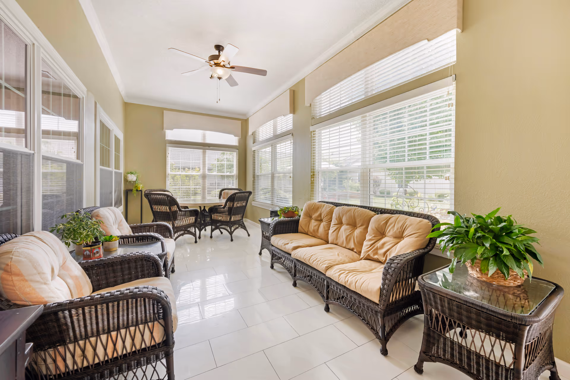 Sunlit enclosed porch with wicker sofas and chairs, potted plants, and large windows.