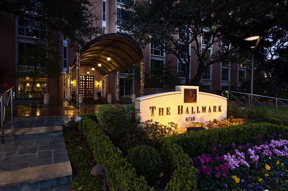 The Hallmark building entrance at dusk with an illuminated sign, covered canopy with string lights, and landscaped flower beds.