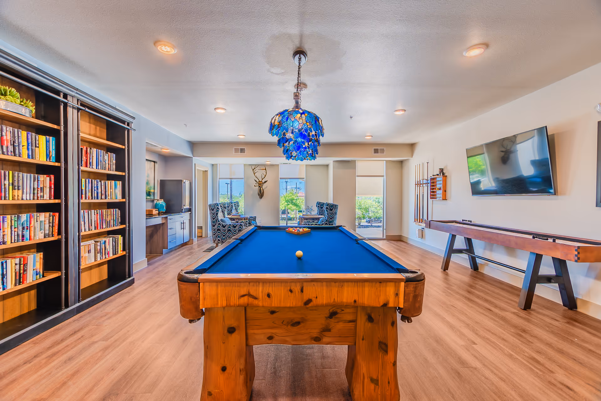 A bright and spacious game room featuring a wooden pool table with blue felt in the center, a blue stained glass chandelier hanging above it, a bookshelf filled with books on the left, two patterned armchairs with a small table near large windows at the back, a mounted deer head on the wall, a shuffleboard table on the right, and a flat-screen TV mounted on the wall.