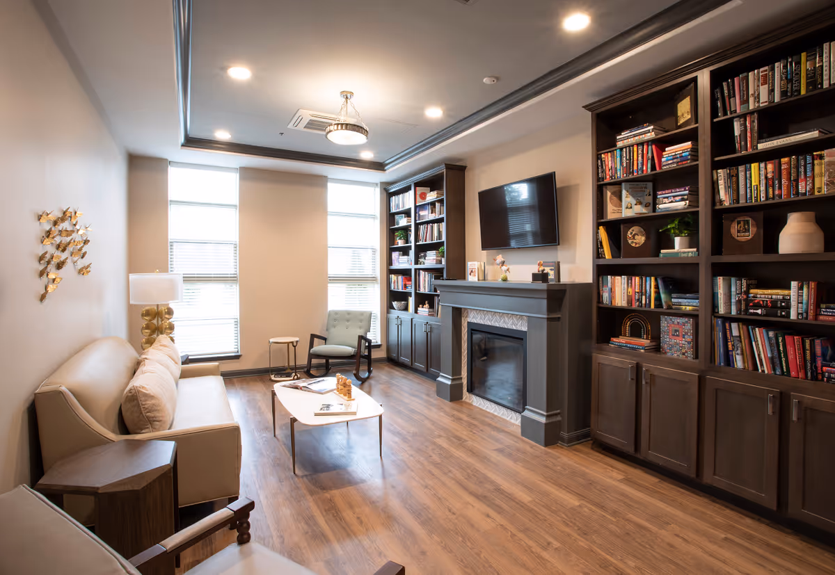 A cozy living room with wooden flooring, a beige sofa with cushions, a small wooden side table, a modern armchair, and a coffee table with books and a chess set. The room features large windows with blinds, built-in dark wood bookshelves filled with books and decorative items, a fireplace with a mounted flat-screen TV above it, and a ceiling light fixture.