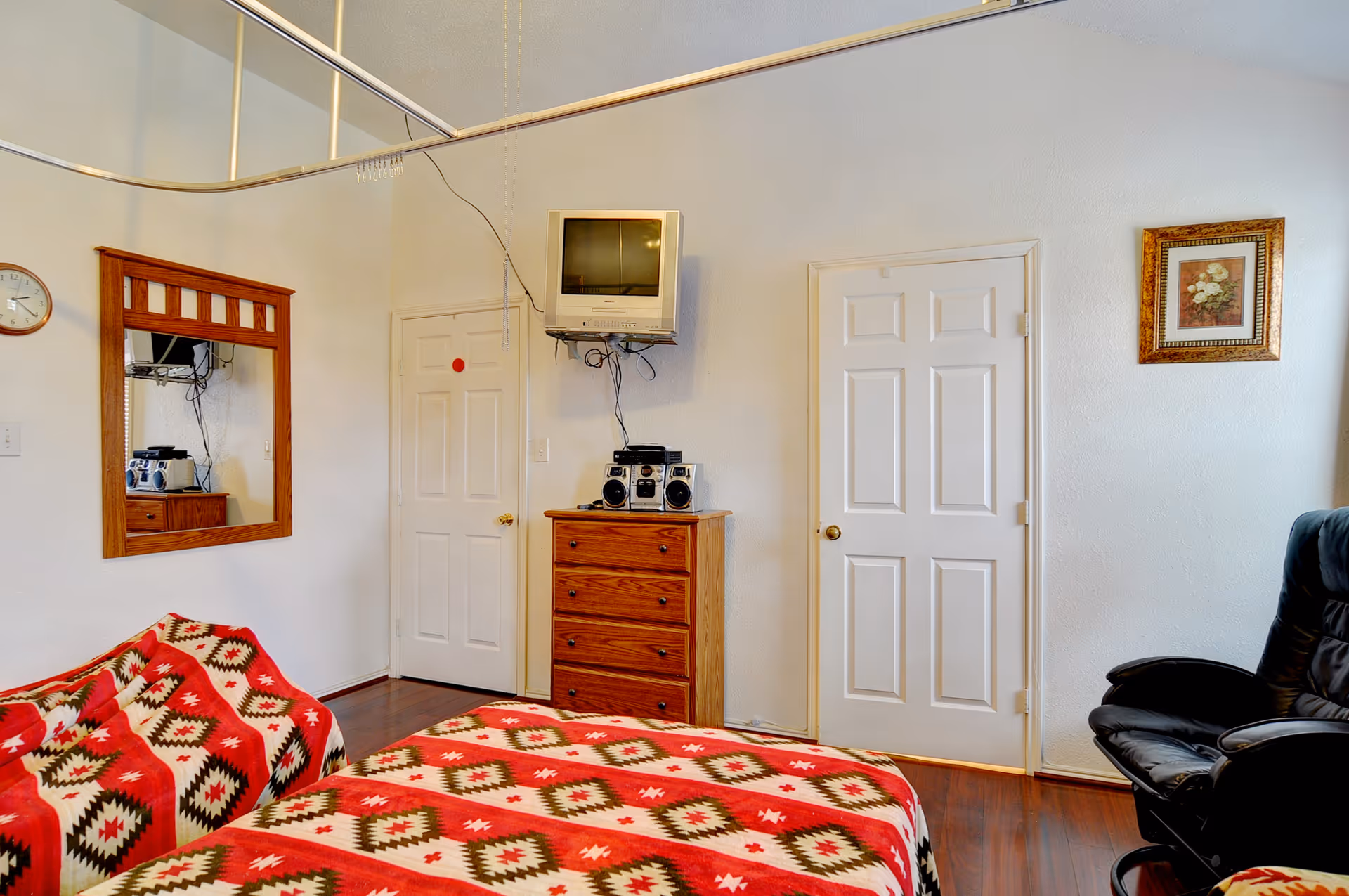 A bedroom with a bed covered in a red, white, and brown patterned blanket. There is a matching armchair to the left, a wooden dresser with a stereo system on top, and a small wall-mounted TV above it. Two white doors are visible on the far wall, along with a framed floral picture and a wooden-framed mirror on the left wall. The floor is wooden, and there is a black leather chair on the right side.