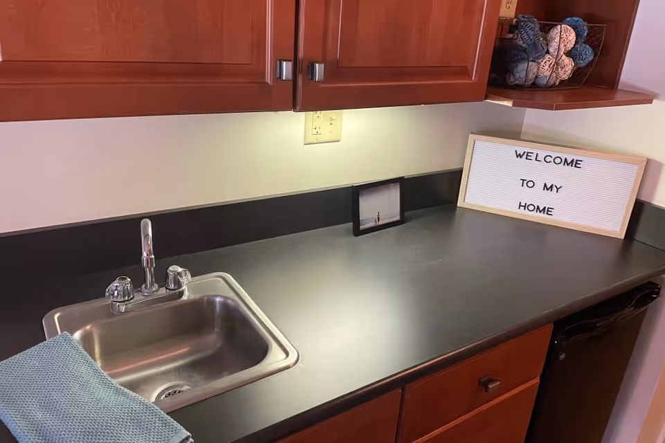 Kitchen countertop with a stainless steel sink and faucet, wooden cabinets above, and a framed sign on the counter reading 'WELCOME TO MY HOME'.