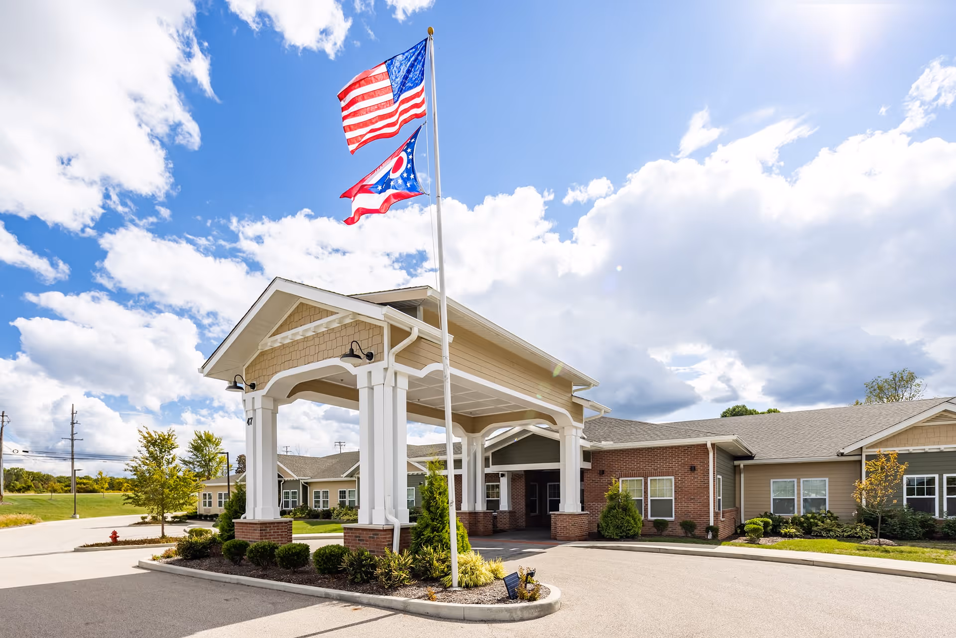 Exterior view of Mulberry Gardens Memory Care facility showing a covered entrance with white columns and brick accents. Two flags, the American flag and the Ohio state flag, are flying on a flagpole in front of the entrance. The sky is partly cloudy with bright sunlight.
