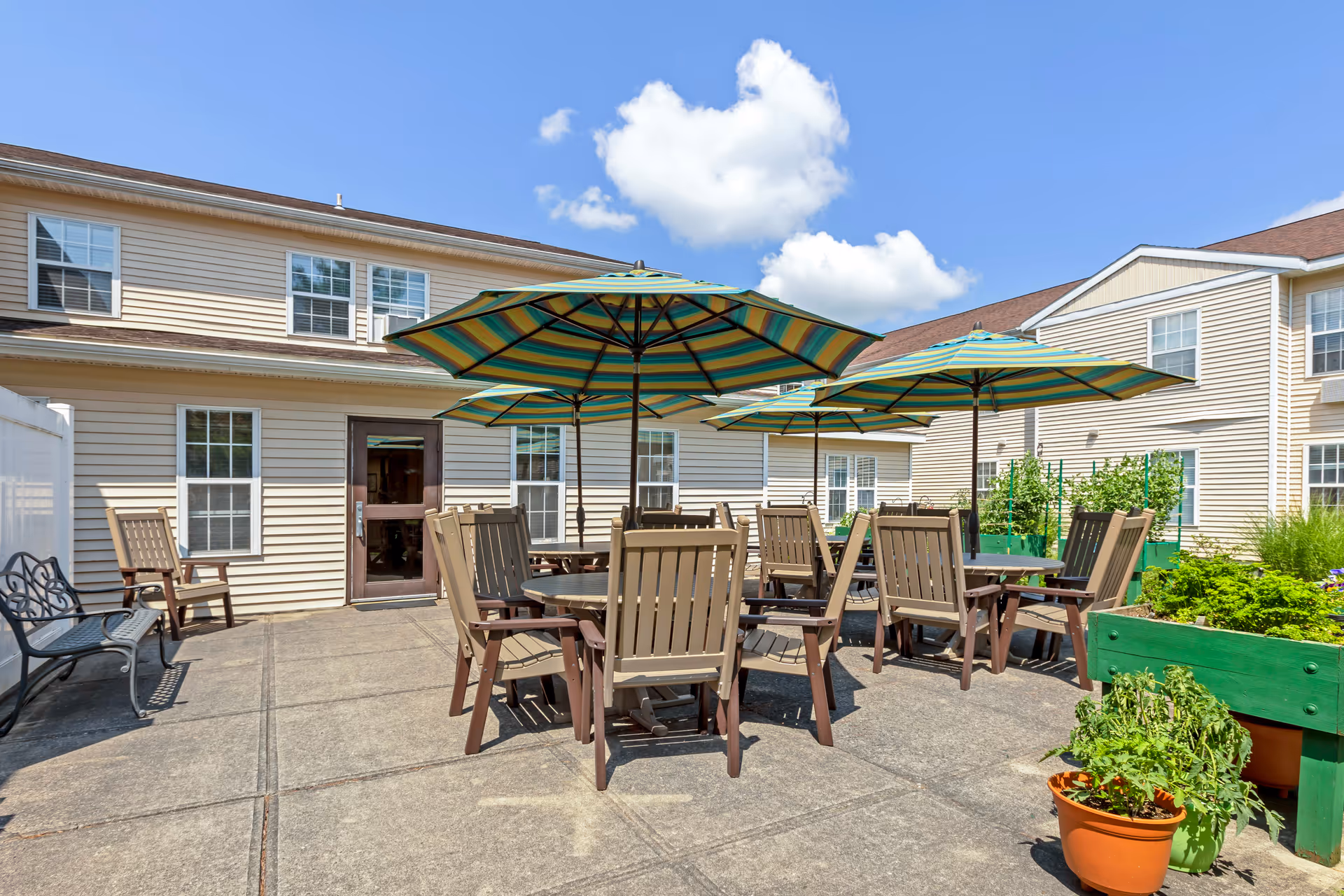 Outdoor patio area at Brookdale Clinton IL with several tables and chairs under large striped umbrellas. The patio is surrounded by beige buildings with multiple windows, and there are green planters and potted plants along the edges. The sky is clear with a few white clouds.