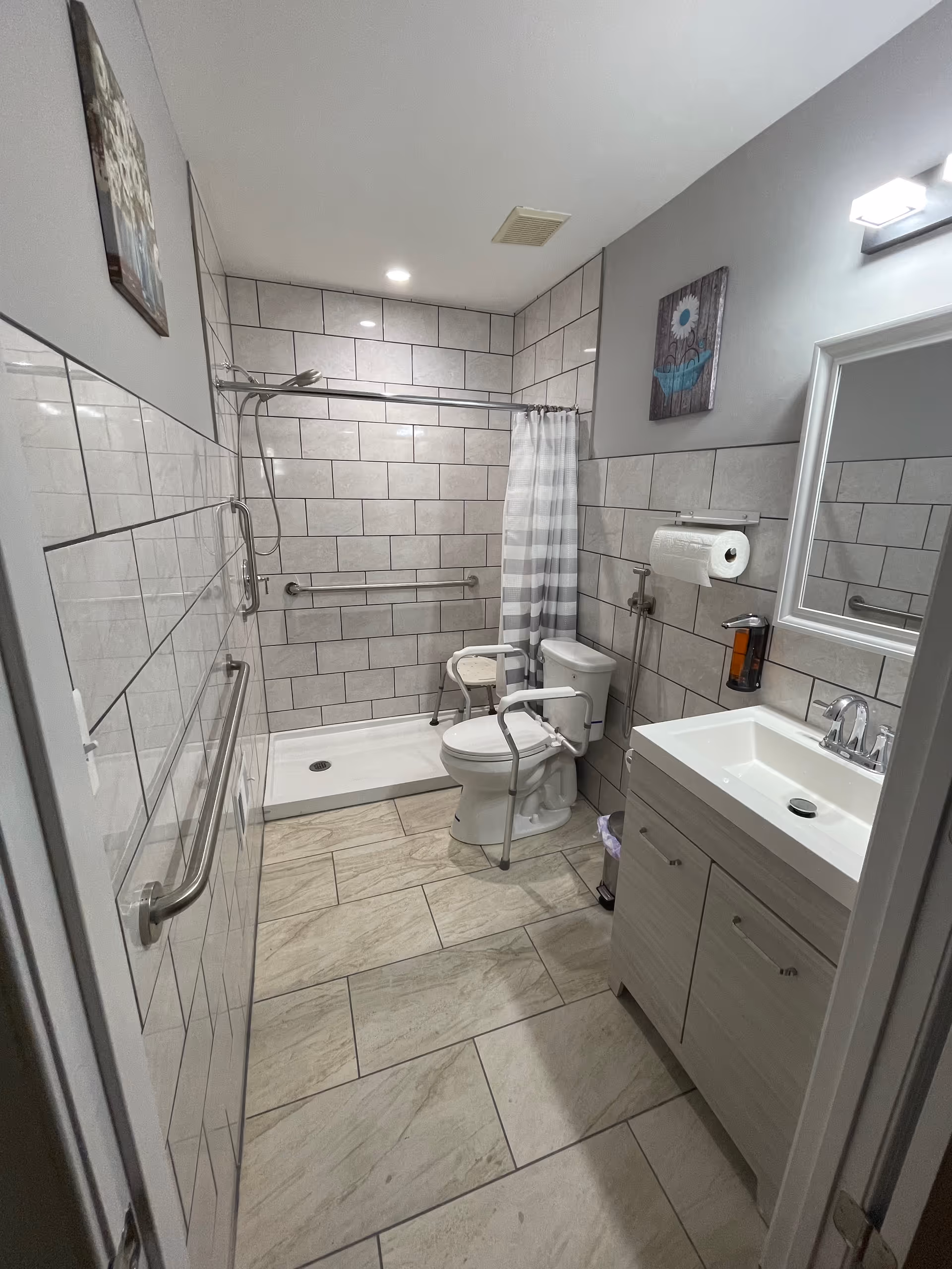 A clean, accessible bathroom with beige tiled walls and floor. The room features a walk-in shower with a white and gray striped shower curtain, a white toilet with safety rails, a white sink with a cabinet below, a mirror above the sink, and handrails on the walls for support. There are two pieces of wall art and a mounted paper towel holder next to the sink.