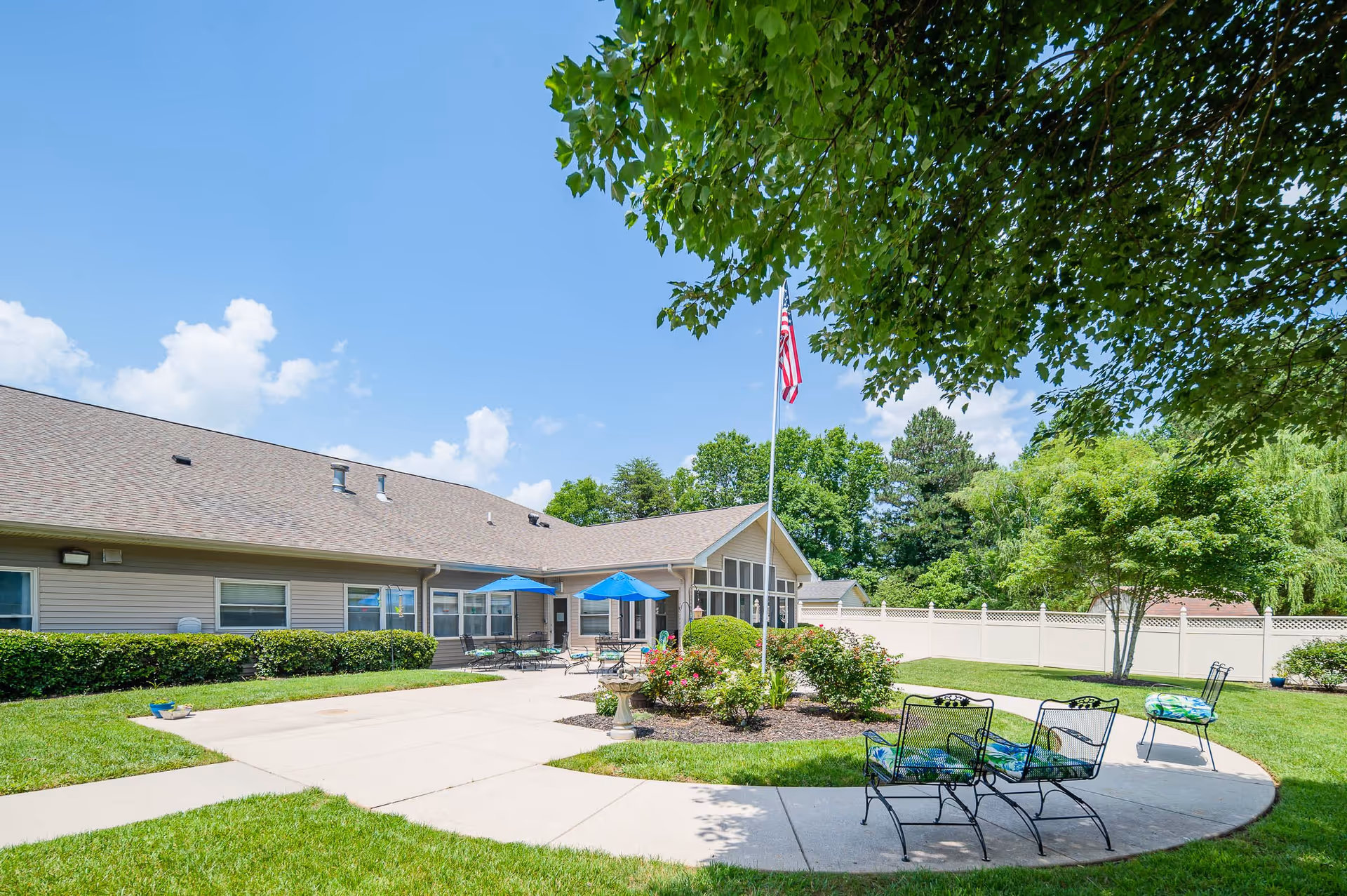 Outdoor patio area at Ascension Living Valley Residence with metal chairs featuring colorful cushions, a concrete walkway, green grass, bushes, and trees. The building has a beige exterior with multiple windows and blue umbrellas shading tables. An American flag is flying on a flagpole near the center of the image under a clear blue sky.