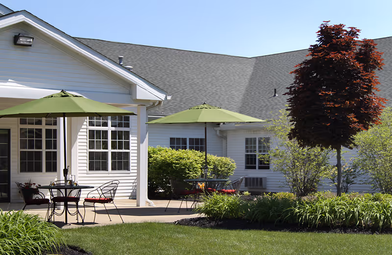 Outdoor patio area at Northfield Village Skilled Nursing & Rehabilitation with green umbrellas shading metal tables and chairs with red cushions, surrounded by green bushes, a red-leafed tree, and a well-maintained lawn in front of a white building with multiple windows.