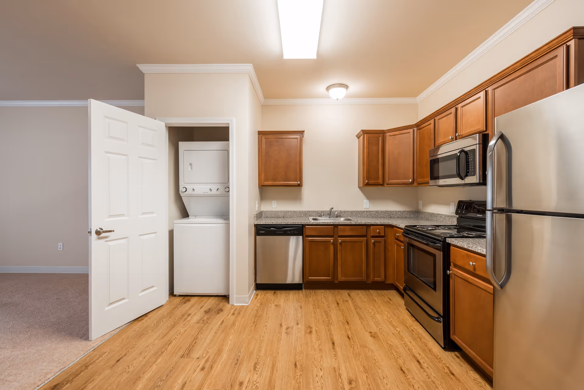 A modern kitchen with wooden cabinets, granite countertops, and stainless steel appliances including a refrigerator, microwave, stove, and dishwasher. To the left, there is an open door revealing a stacked washer and dryer unit. The floor is wood, and the walls are painted a light beige color.