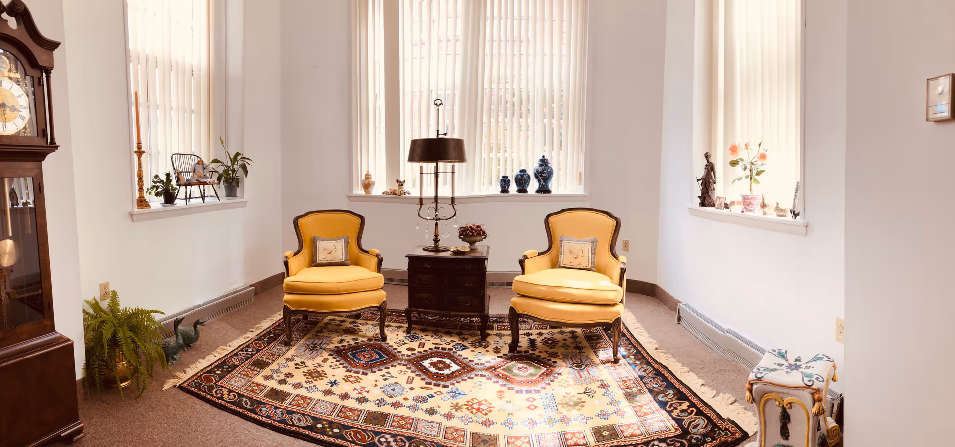 A cozy sitting area with two yellow upholstered armchairs placed on a patterned rug. Between the chairs is a dark wooden side table with a lamp and decorative items. The room has three tall windows with vertical blinds, and various decorative objects and plants are placed on the windowsills. A grandfather clock is visible on the left side, and a small decorative stool is on the right.