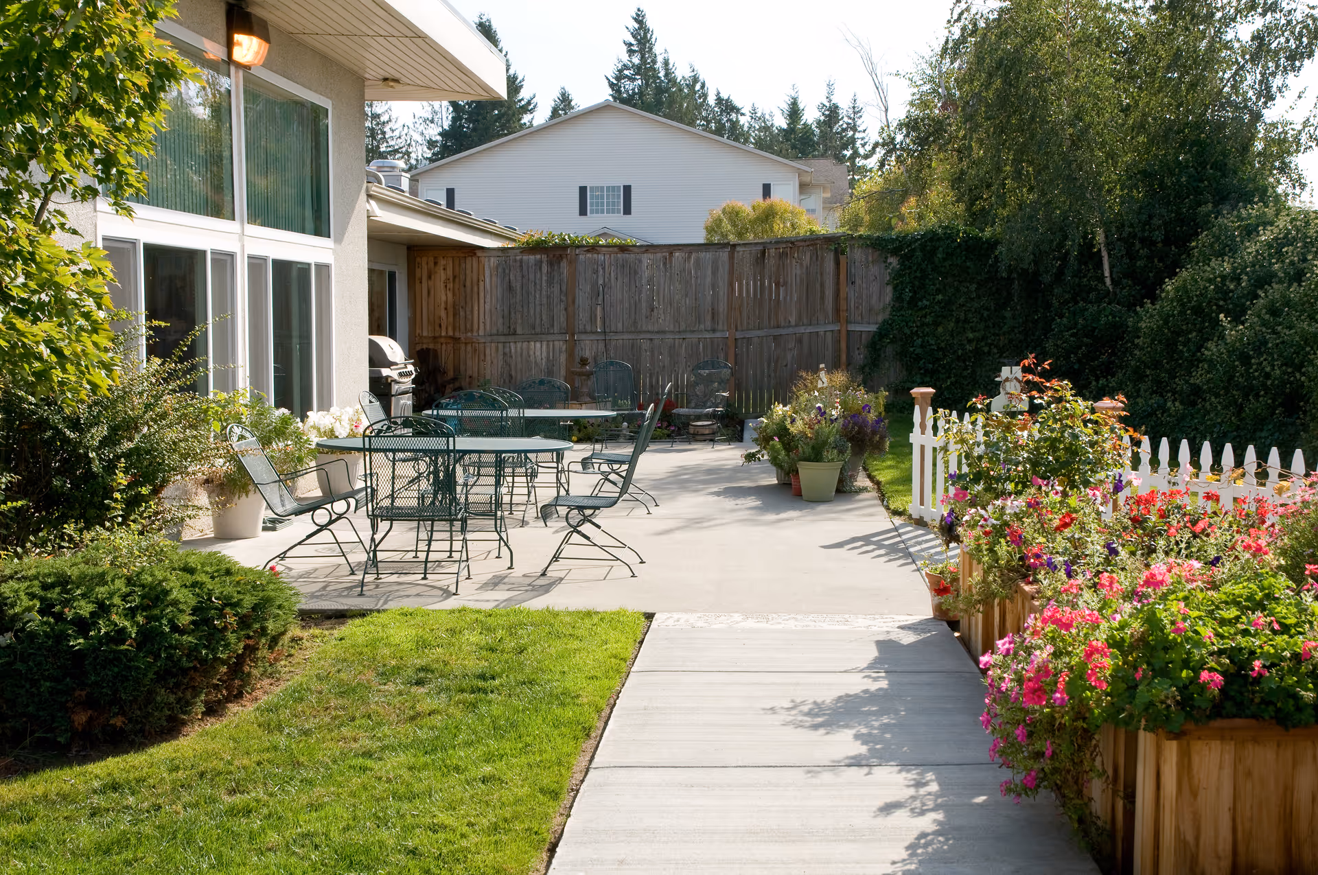 Outdoor patio area at a senior living facility with metal tables and chairs on a concrete surface, surrounded by green bushes, colorful flowers in wooden planters, and a white picket fence. The building has large windows and a barbecue grill is visible near the wall. Trees and a wooden fence are in the background.