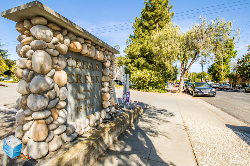 Stone sign for Cedar Creek Alzheimer's and Dementia Care located next to a sidewalk with trees and parked cars along the street on a sunny day.
