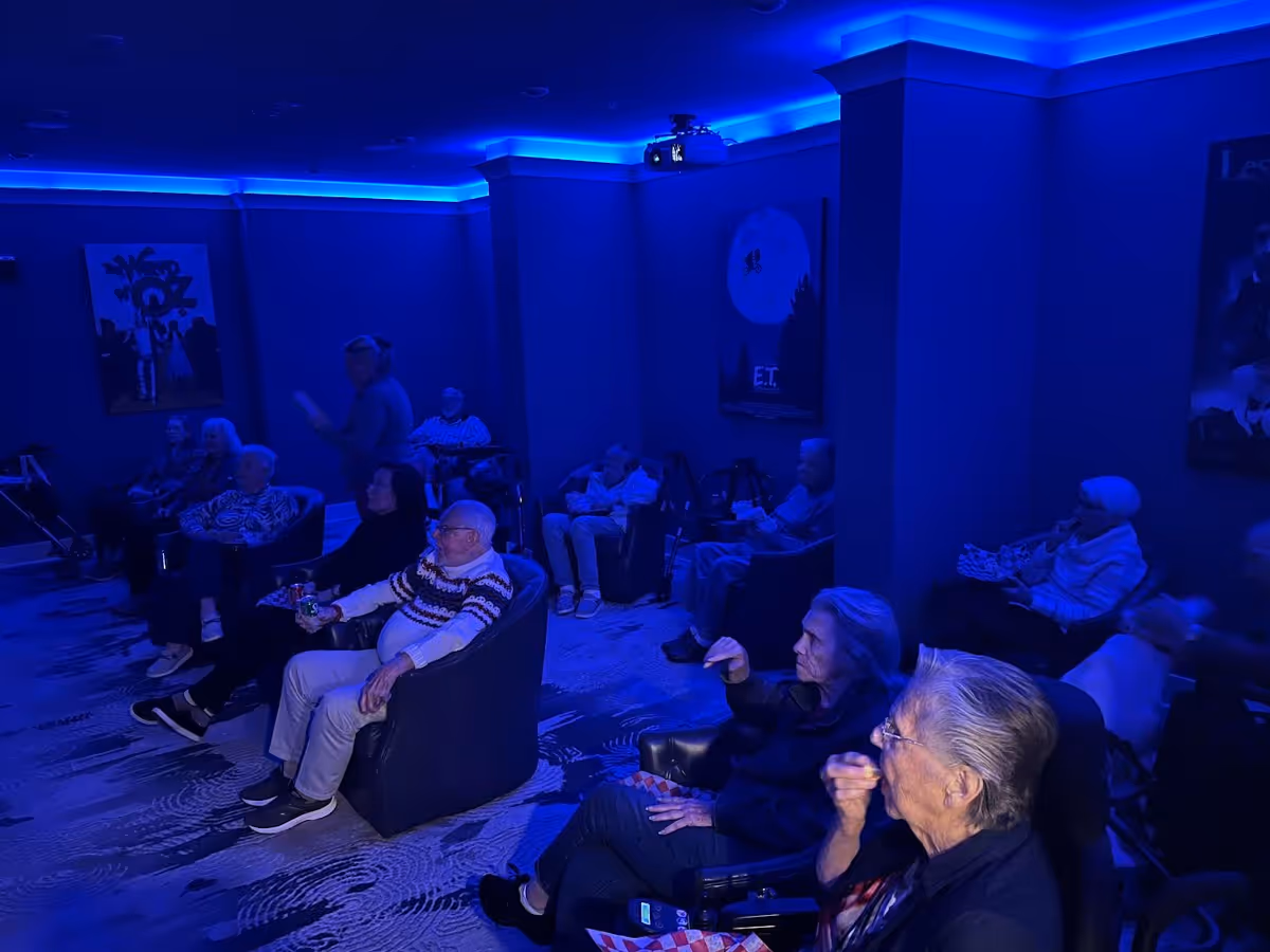 A group of elderly people seated in a dimly lit room with blue lighting, watching a movie or presentation. The room has movie posters on the walls and comfortable chairs arranged in rows.