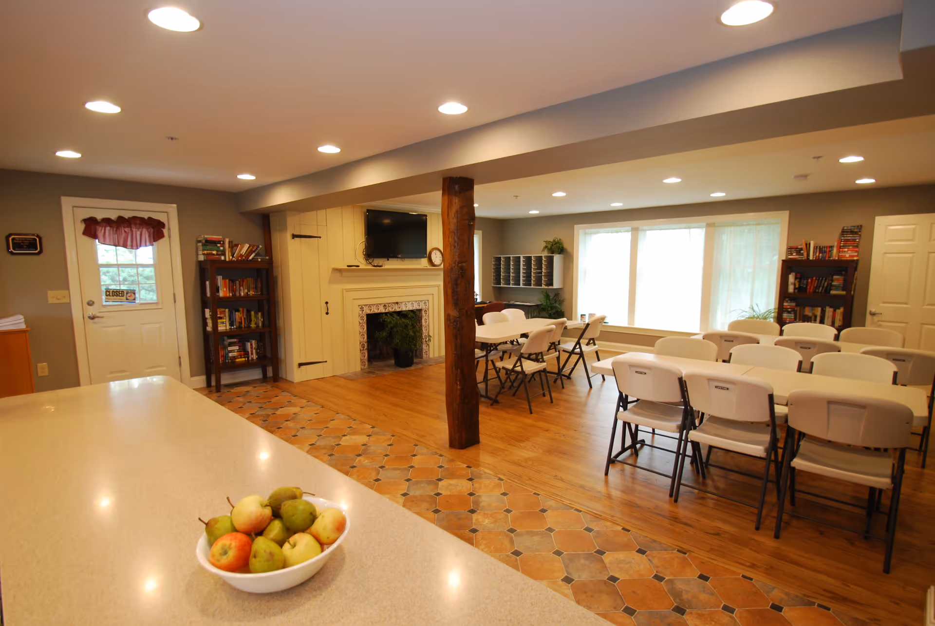 A spacious community room with a long countertop in the foreground holding a bowl of apples and pears. The room features a wooden floor with a tiled border, several folding tables and chairs arranged for group seating, bookshelves filled with books, a fireplace with a mounted TV above it, and large windows letting in natural light.