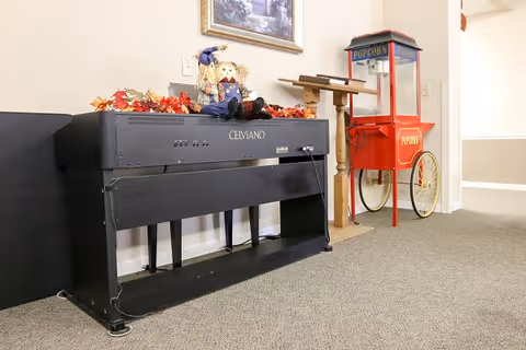 A black Celviano digital piano against a beige wall with autumn-themed decorations and a small scarecrow doll on top. Next to the piano is a vintage-style red popcorn machine with yellow wheels. The floor is carpeted and there is a wooden stand behind the popcorn machine.