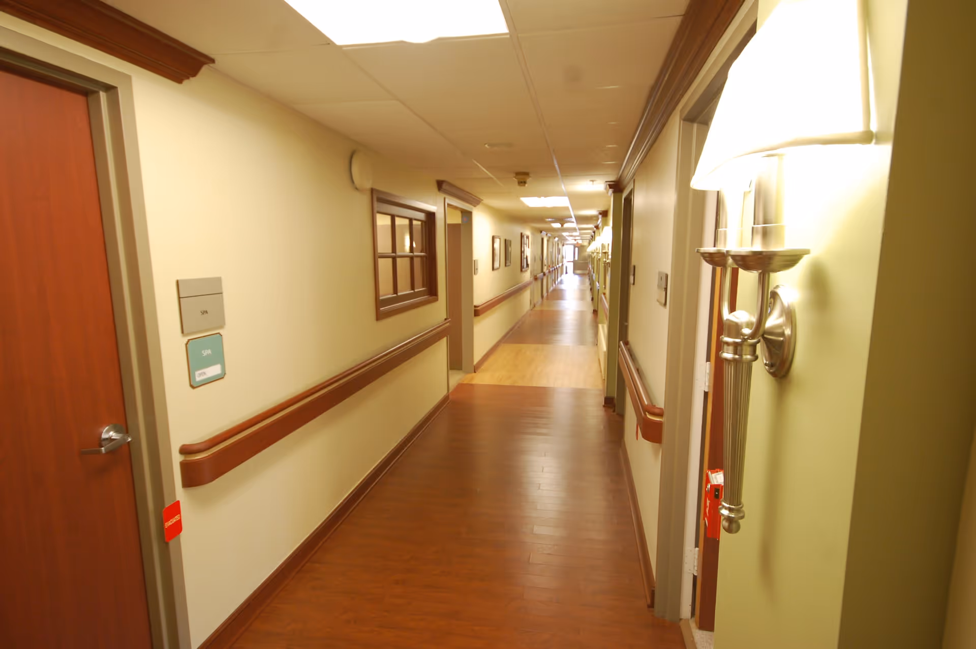 A long, well-lit hallway in a senior living facility with wooden floors and handrails on both sides. Doors and wall-mounted lamps line the corridor, and there are framed pictures on the walls. The ceiling has recessed lighting panels.