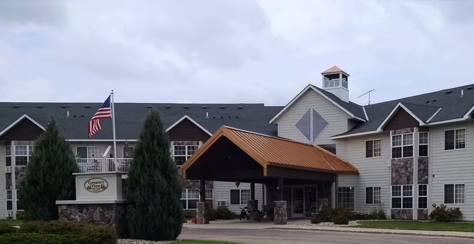 Exterior front view of a senior living facility building with a covered entrance, stone and siding facade, multiple windows, and an American flag on a flagpole near the entrance sign that reads 'Country View Senior Living'.