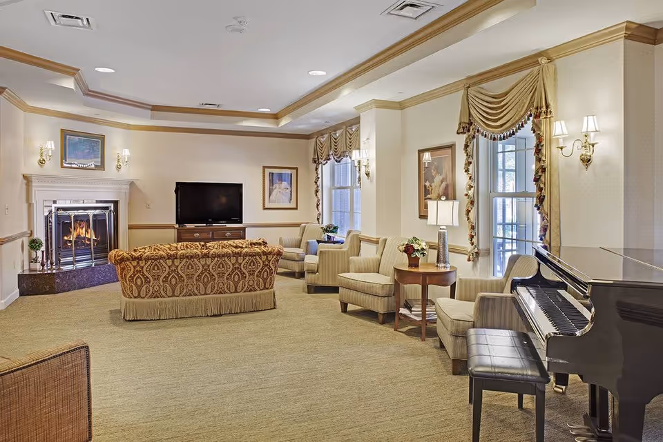 A cozy living room with a patterned sofa facing a fireplace and a flat-screen TV. The room has beige walls with crown molding, several armchairs, a side table with a lamp and flowers, large windows with draped curtains, and a black grand piano with a bench.