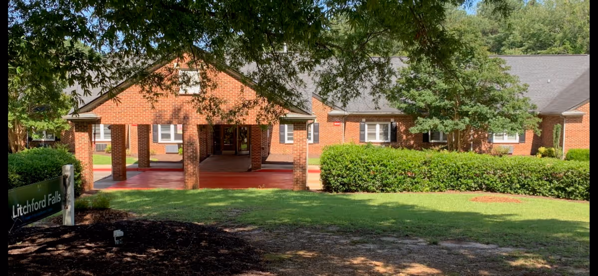 Exterior view of Litchford Falls Health & Rehabilitation Center showing a brick building with a covered entrance, surrounded by green bushes and trees under a clear sky.