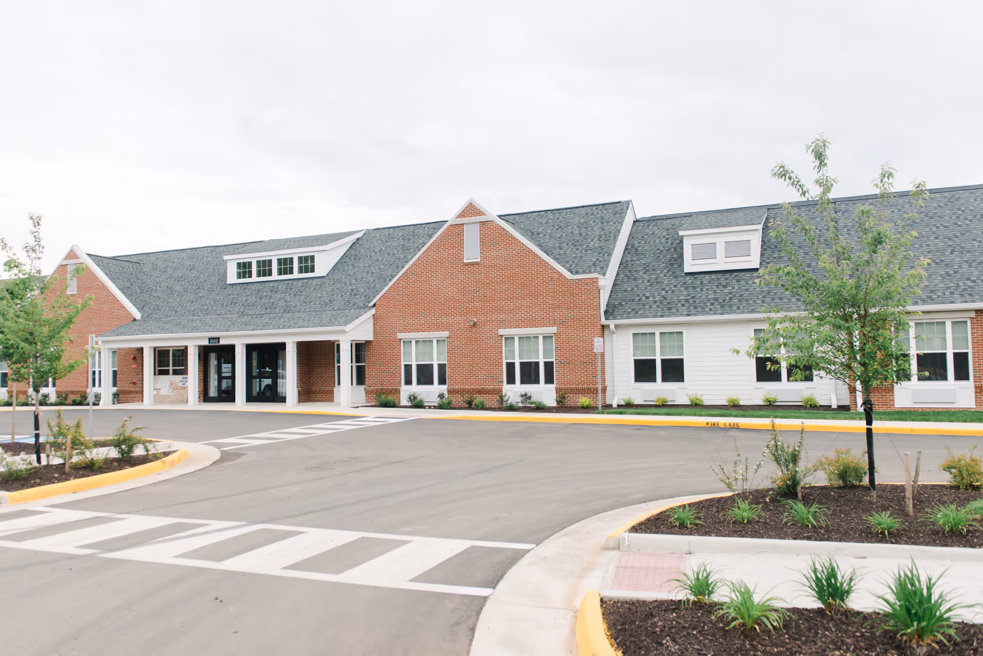 Front exterior view of a single-story senior living facility building with a combination of red brick and white siding, gray shingled roof, and multiple windows. There is a driveway and landscaped areas with small trees and plants in front of the building under an overcast sky.