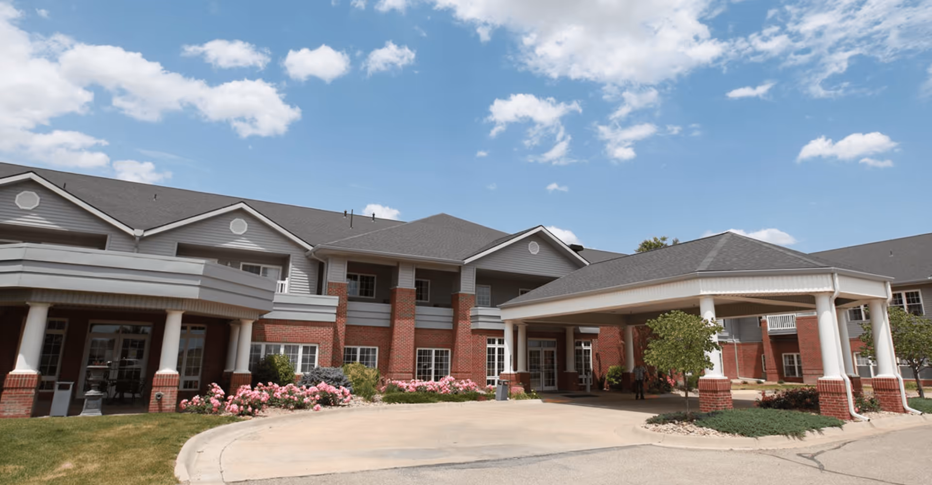 Exterior view of Eaglecrest Senior Living of Salina building with a covered entrance supported by white columns, red brick and gray siding, landscaped with green bushes and pink flowers under a partly cloudy blue sky.