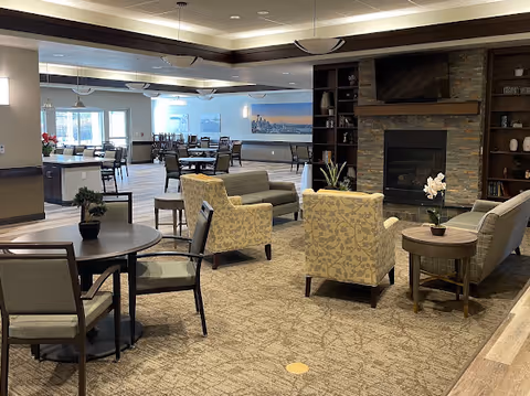A spacious and well-lit common area in a senior living facility featuring multiple seating arrangements including cushioned armchairs, sofas, and round tables with chairs. There is a stone fireplace with a mounted flat-screen TV above it, built-in shelves with decorative items, and large windows letting in natural light. The room has carpeted flooring and modern ceiling lights.