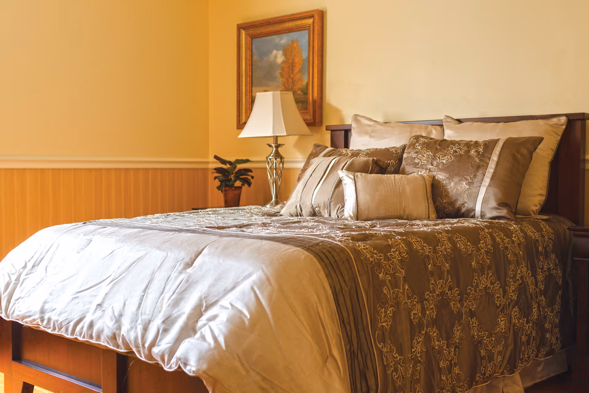 A neatly made bed with multiple decorative pillows and a patterned bedspread in warm brown and beige tones. Next to the bed is a wooden nightstand with a silver lamp and a small potted plant. A framed painting of a tree with autumn leaves hangs on the wall above the nightstand. The walls are painted a soft yellow with wooden paneling on the lower half.