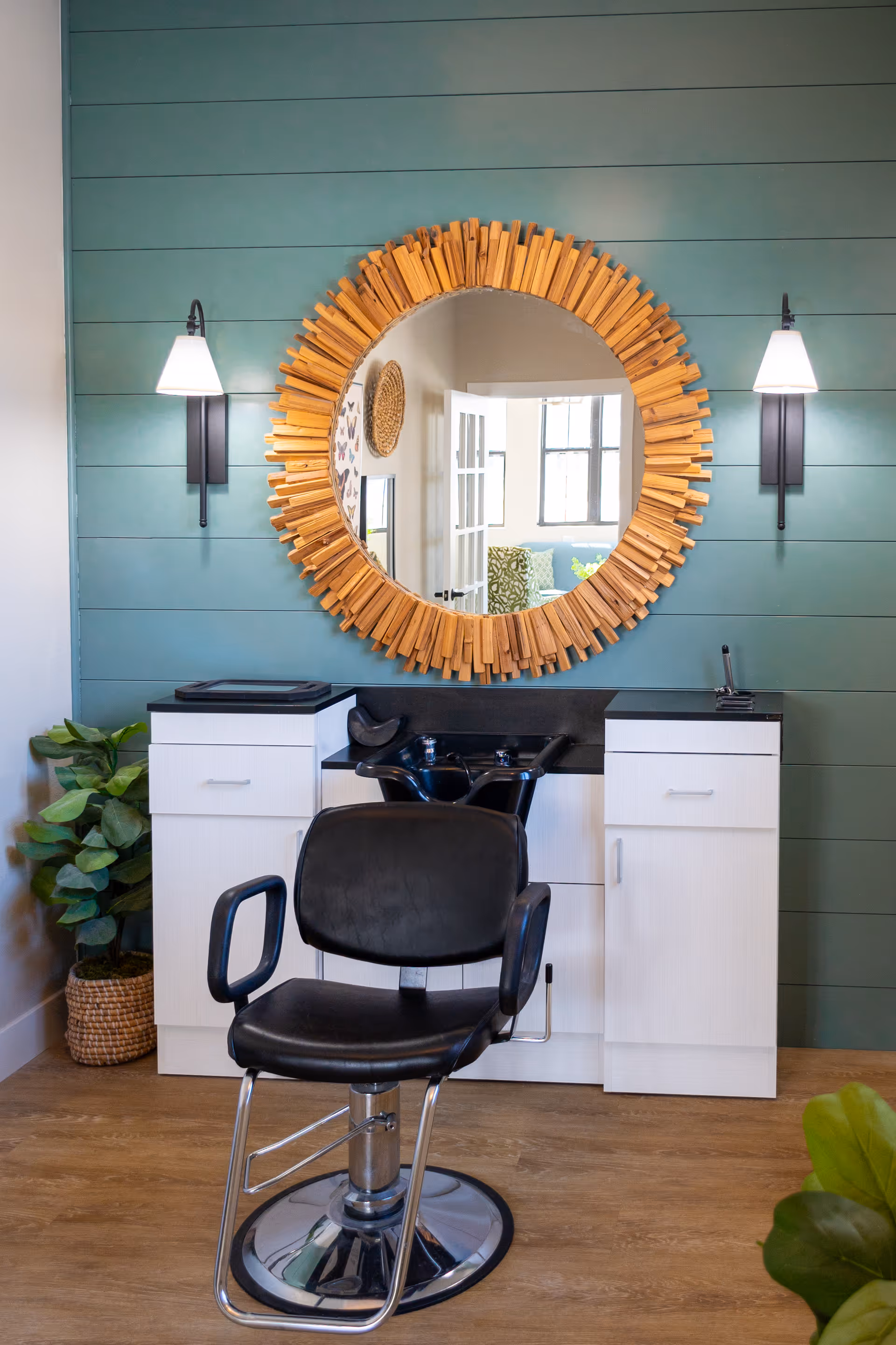 A salon area with a black salon chair in front of a white cabinet with a built-in black sink. Above the sink is a large round mirror with a decorative wooden frame. The wall behind is painted teal with horizontal panels and has two wall-mounted lights on either side of the mirror. There is a potted plant on the left side of the cabinet.