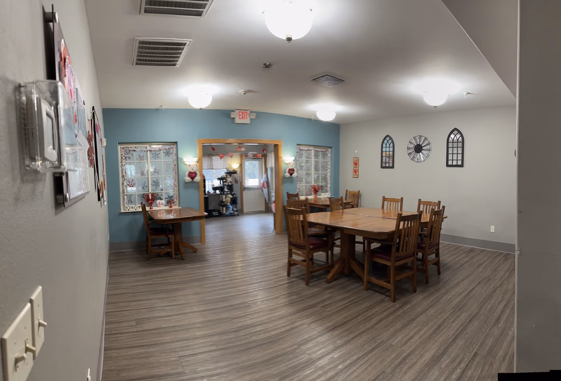 Communal dining area with wooden tables and chairs, wall decor, and a view into an adjacent room.