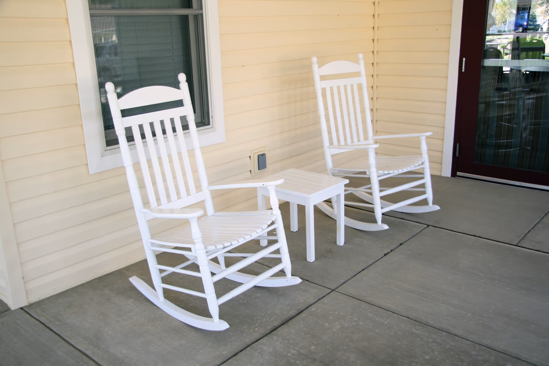 Two white wooden rocking chairs with a small matching white table between them on a concrete porch outside a building with beige siding and a window with closed blinds.