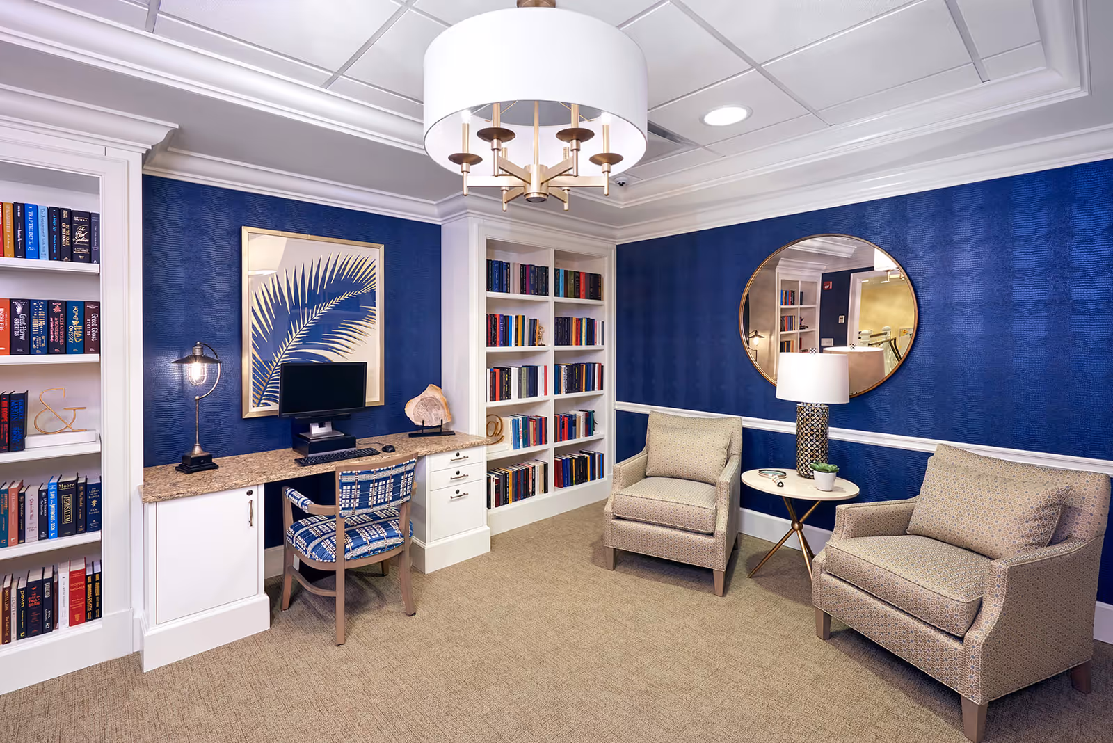 A cozy reading and sitting area with blue textured walls, beige carpet, and white trim. The room features a built-in bookshelf filled with books, a desk with a computer and a patterned chair, two beige armchairs with a small round table and lamp between them, a large round mirror on the wall, and a modern ceiling light fixture.