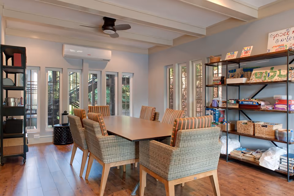 A well-lit room with a rectangular wooden table surrounded by eight wicker chairs with striped cushions. The room has wooden flooring, multiple tall narrow windows letting in natural light, and a wall-mounted air conditioning unit. On the right side, there is a black metal shelving unit filled with various board games, books, baskets, and craft supplies. Another shelving unit is visible on the left side of the room.