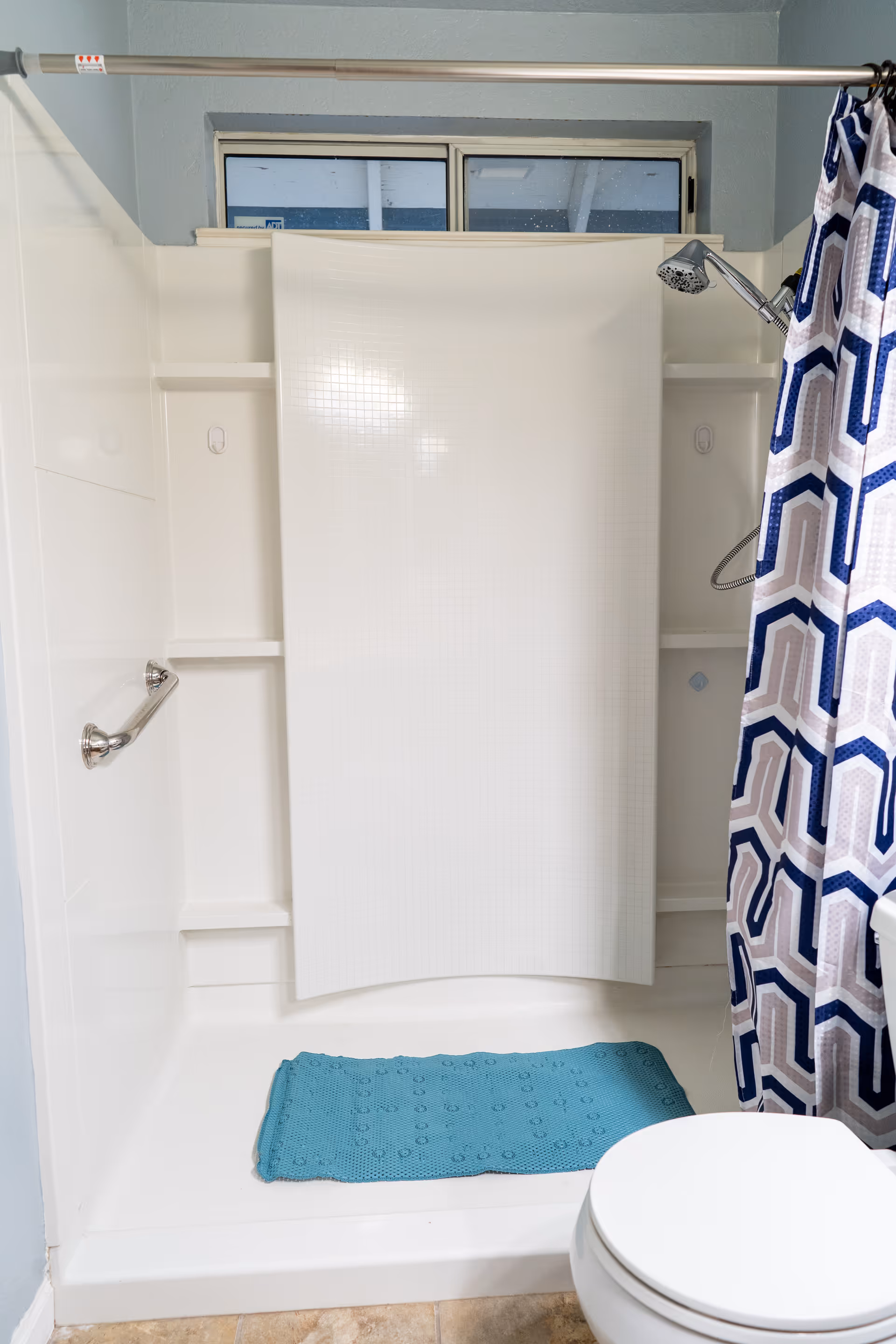 A bathroom shower stall with a white surround, chrome grab bar and showerhead, a blue geometric shower curtain, teal bath mat, and part of a toilet visible.
