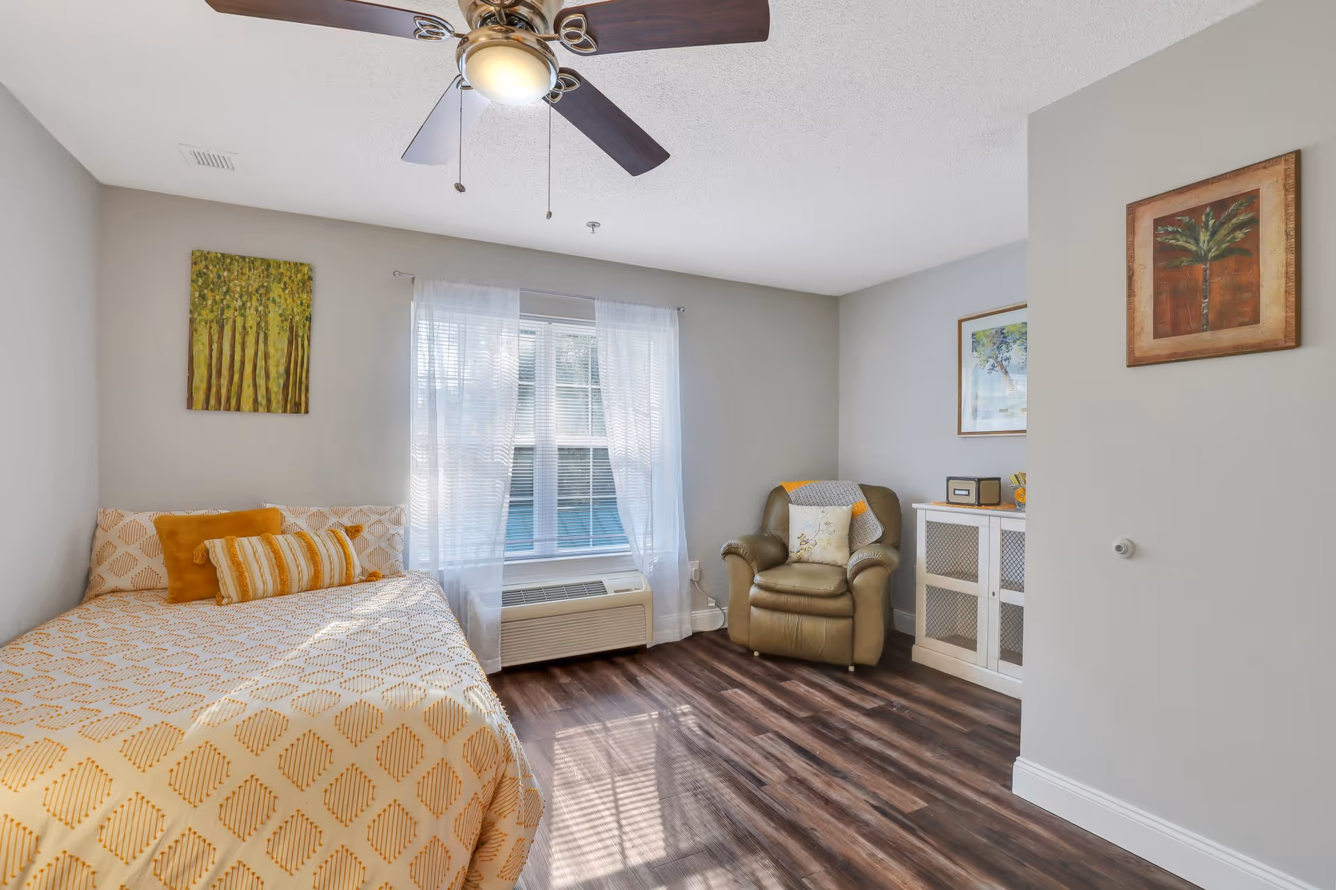 A bright and cozy bedroom with a bed covered in a yellow patterned bedspread and matching pillows, a large window with sheer white curtains, a ceiling fan with lights, a comfortable armchair with a throw blanket and pillow, a white cabinet with decorative items, and framed artwork on the walls.