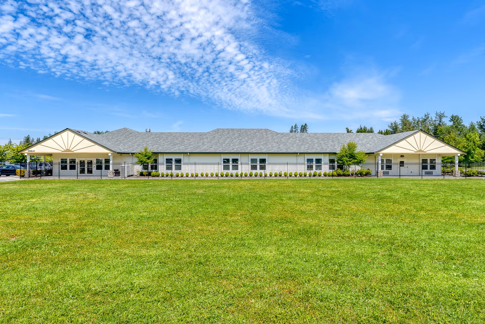 Wide exterior view of a single-story assisted living facility building with a gray roof, white walls, and multiple windows. The building is surrounded by a black metal fence and small shrubs, with a large green lawn in the foreground and a blue sky with scattered clouds above.