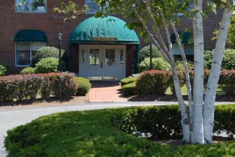 Brick building entrance with a green awning and manicured shrubs.