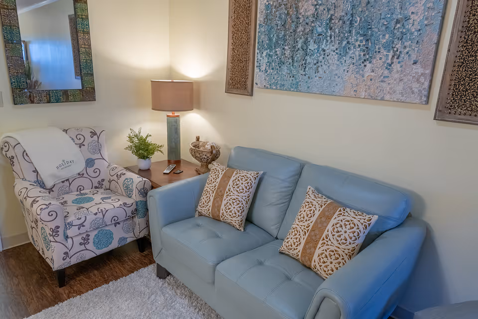 A cozy living room area with a light blue leather sofa adorned with two decorative pillows featuring intricate patterns. Next to the sofa is a wooden side table with a green ceramic lamp, a small potted plant, and a decorative bowl. To the left, there is a patterned armchair with a white throw blanket draped over the back. The walls are decorated with a large abstract painting and two carved wooden panels. A mirror with a decorative frame hangs on the adjacent wall, and the floor is covered with a white rug and wood flooring.