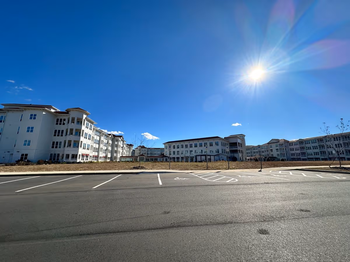 Wide exterior view of a large white multi-story senior living building under a bright sun with an empty parking lot in front.