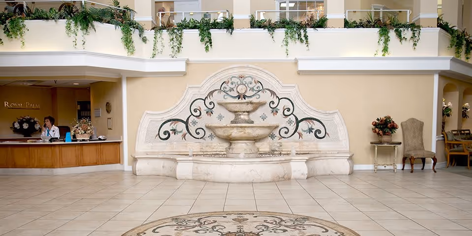 Spacious and elegant lobby area of the Royal Palms facility featuring a decorative stone fountain with floral and scroll designs on the wall behind it. To the left, there is a reception desk with a staff member seated, and to the right, there are chairs and a small table with a flower arrangement. The upper level has a balcony with green plants hanging over the railing.