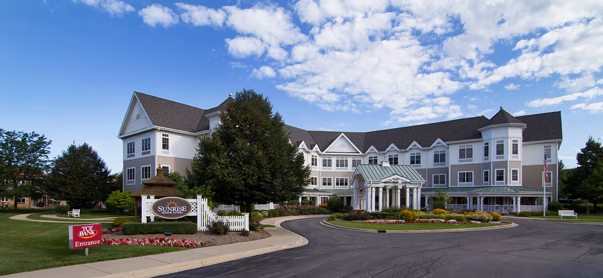Front exterior of Sunrise of Naperville North, a three-story senior living building with a circular driveway, landscaped grounds, and a blue sky.