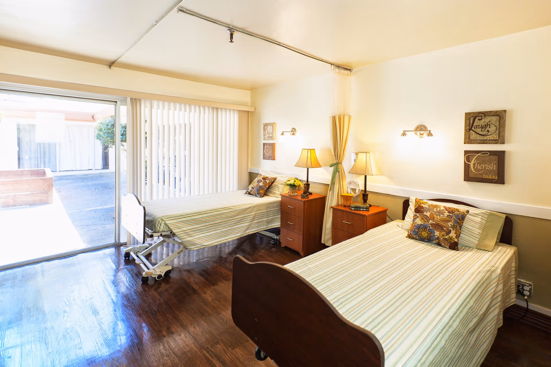 A bright and clean bedroom in a healthcare facility featuring two single beds with striped bedding and decorative pillows. Each bed has a wooden nightstand with a lamp and small decor items. The room has wooden flooring and a large sliding glass door with vertical blinds that opens to an outdoor area. Wall decorations include two framed signs with the words 'Laugh' and 'Cherish'.