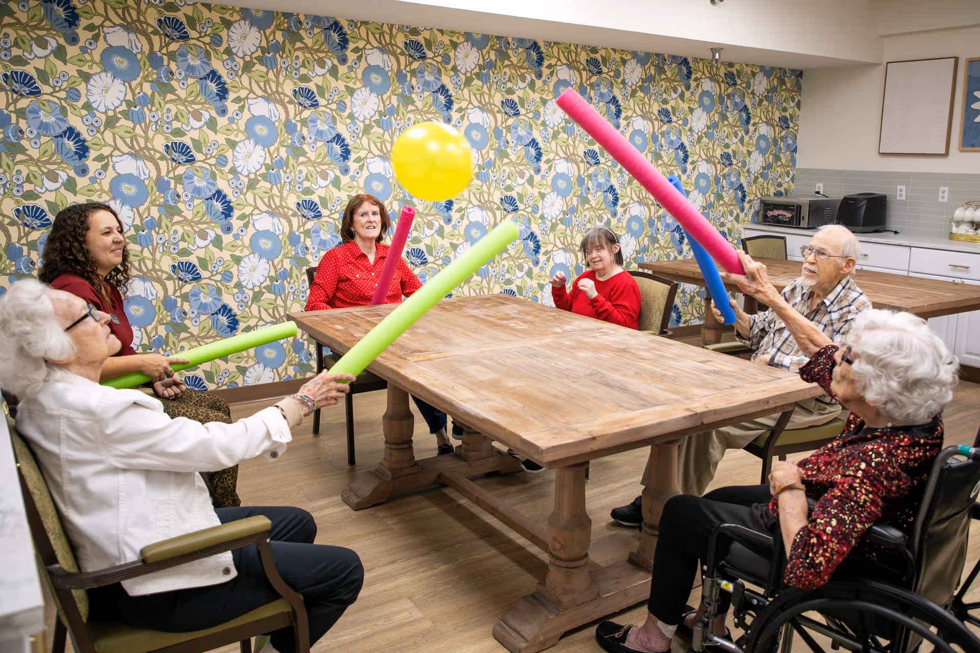 A group of elderly people and a caregiver sitting around a wooden table in a room with floral wallpaper, playing a game with colorful pool noodles and a yellow balloon.
