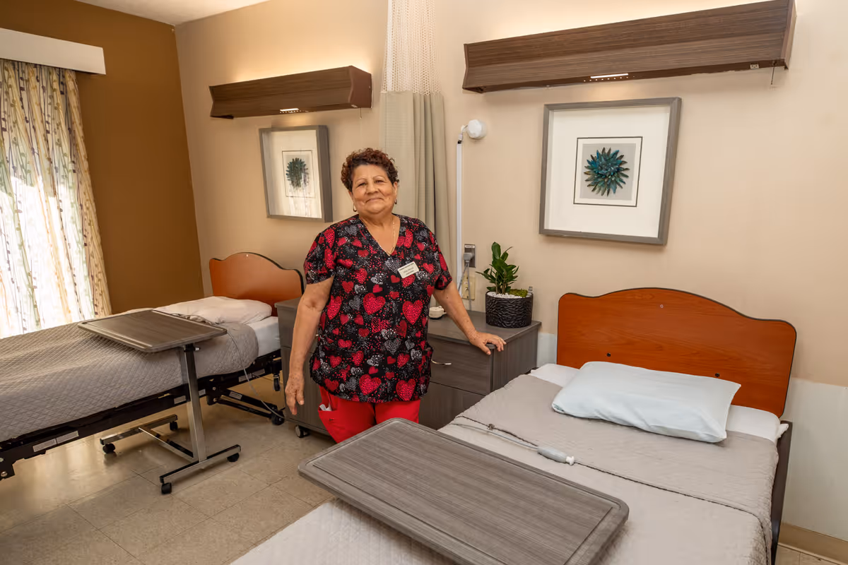 A woman standing in a healthcare facility room with two hospital beds, each with a gray blanket and an overbed table. The room has beige walls, framed artwork, a bedside cabinet with a plant, and a window with patterned curtains.