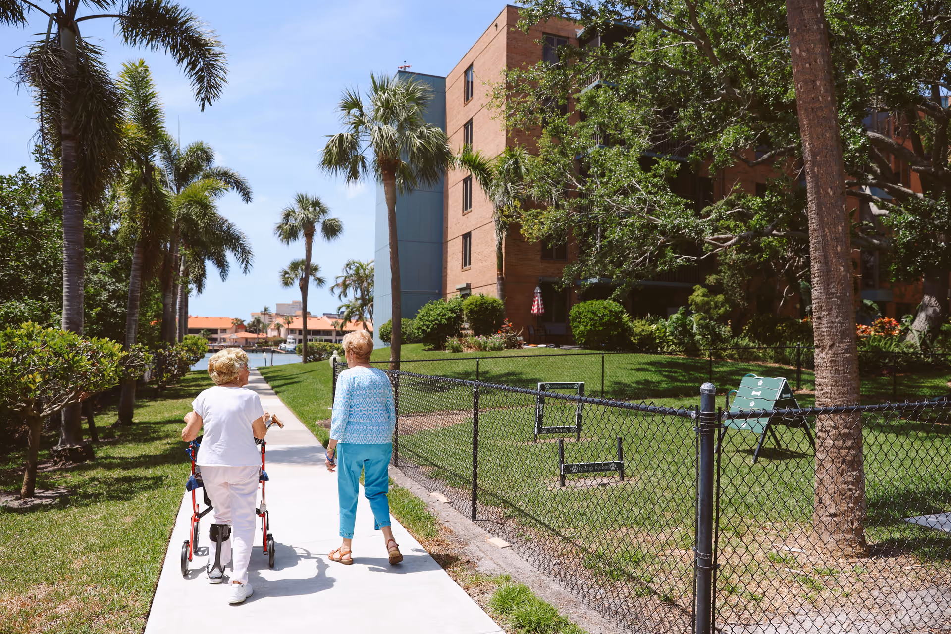 Two elderly women walking on a paved path alongside a fenced grassy area with palm trees and a multi-story brick building in the background under a clear blue sky.